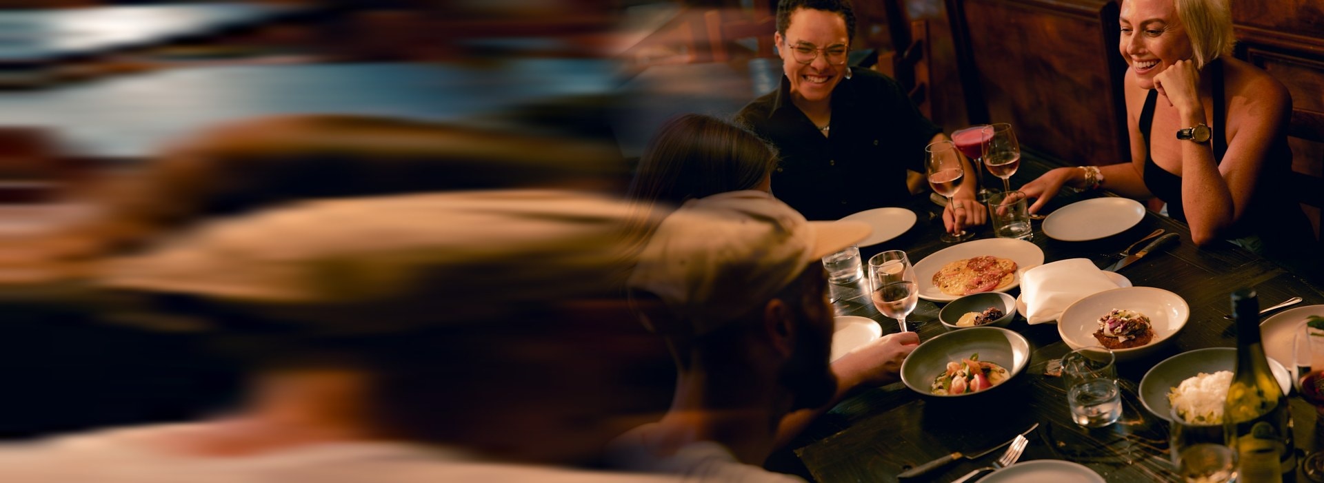 Four people laughing over a shared table of plates and wine in a dark timber room.