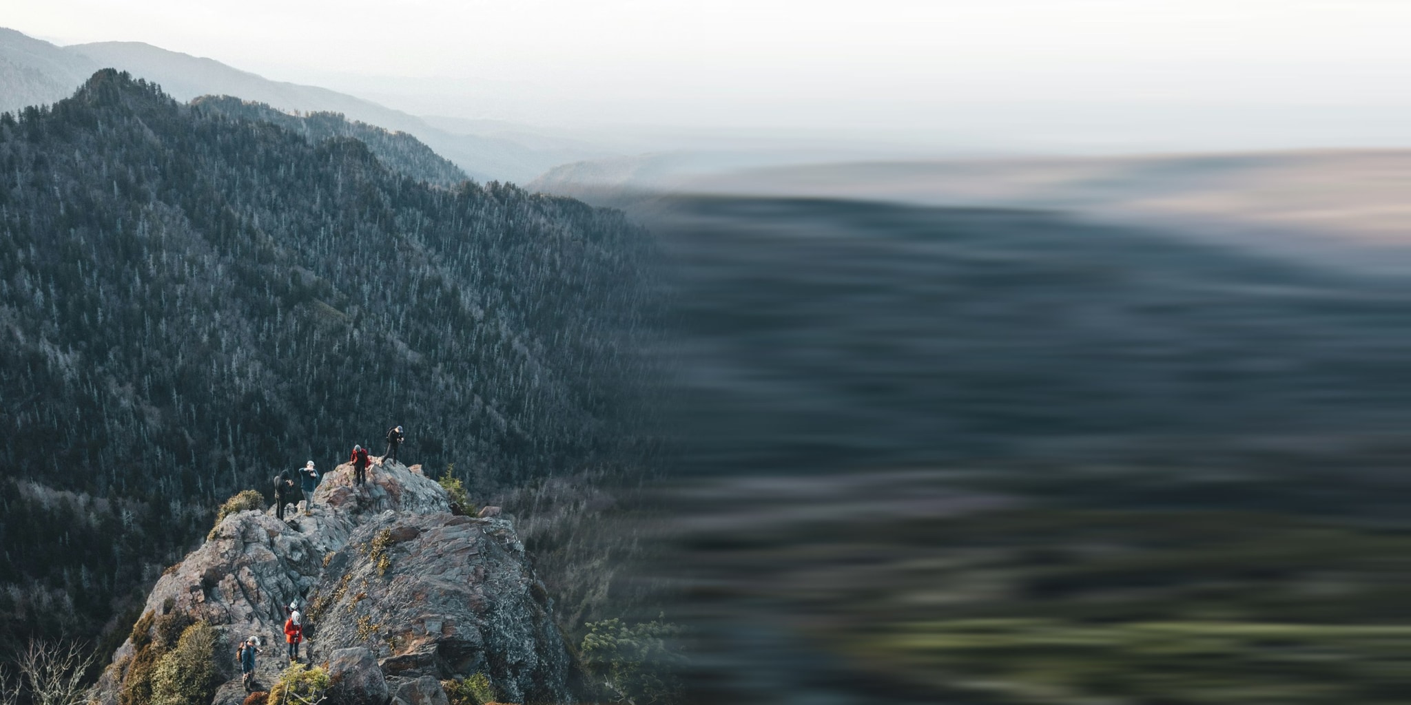 Four people on a rock summit above a forested ridge, late light across the valley behind them.