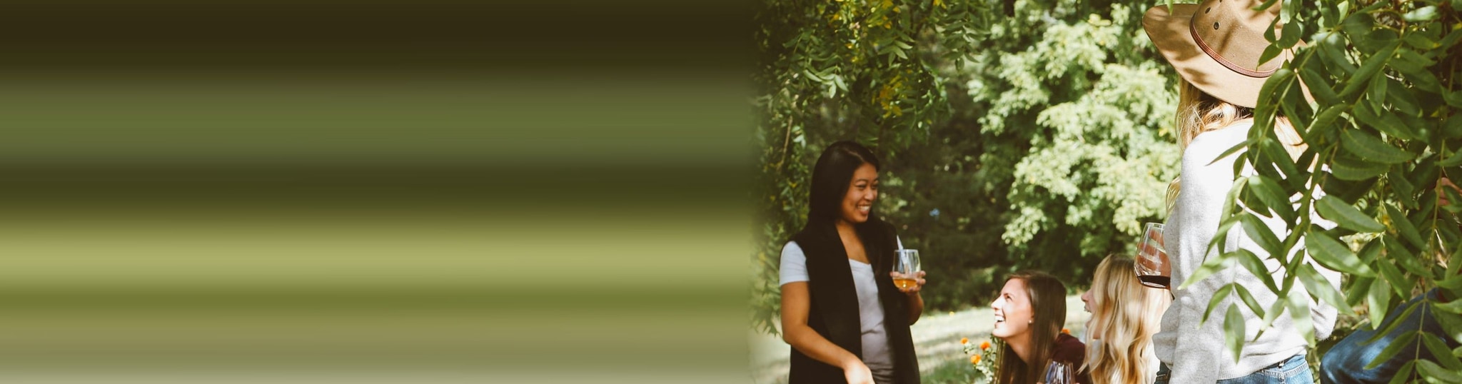 Four women with glasses of wine under the trees on a bright afternoon, one of them standing in a wide-brimmed hat.